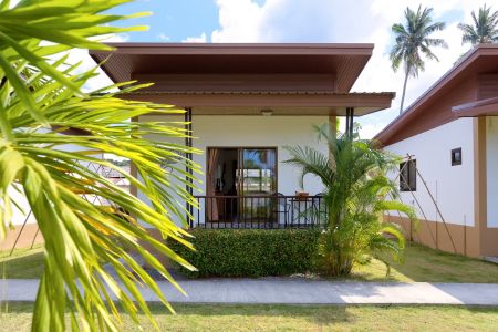 Exterior of a Garden Villa with its covered terrace, hedge and coconut palms behind.